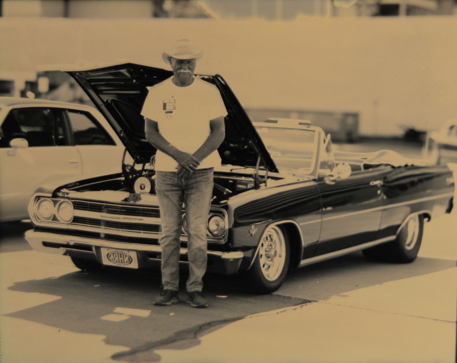 Man in cowboy hat stands proudly beside a classic black convertible muscle car with hood open at an outdoor car show.