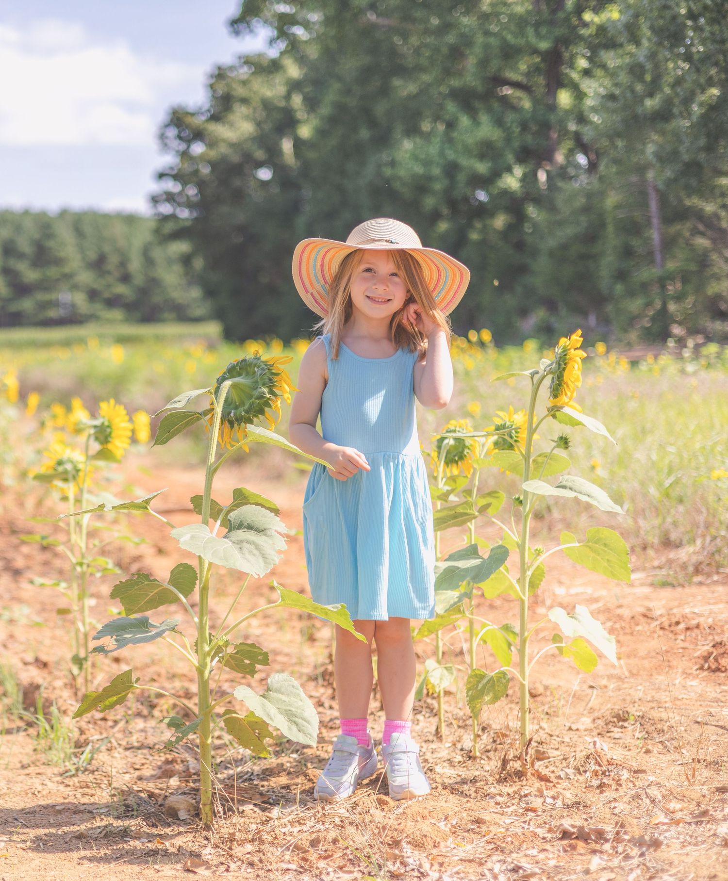 Young girl in light blue dress and sun hat smiling among sunflowers in a sunny field.