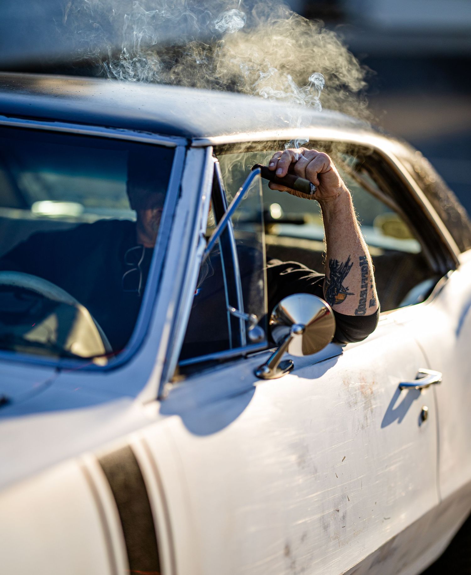 Smoke drifts from a tattooed arm resting on the window frame of a classic white car.