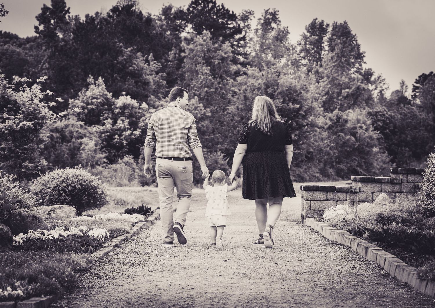 Black and white photo of a family walking hand in hand down a garden path surrounded by lush trees.