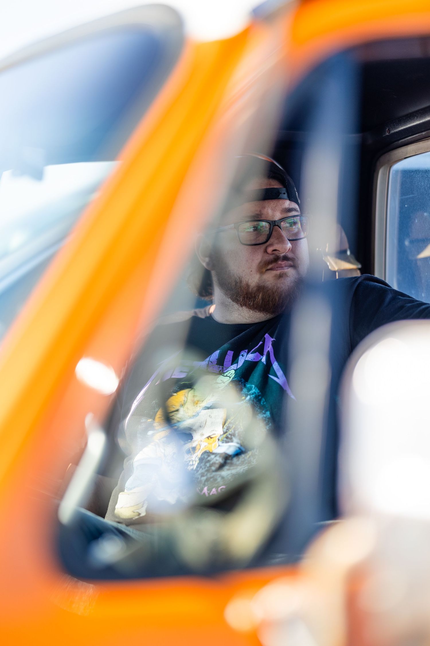 A bearded man wearing glasses sits inside an orange truck, viewed through the window reflection.