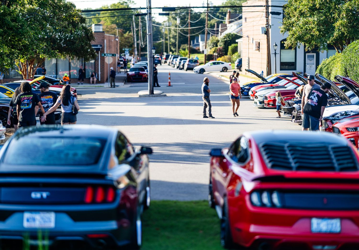 Ford Mustang GT cars lined up on a sunny street during an outdoor car show event with spectators walking nearby.