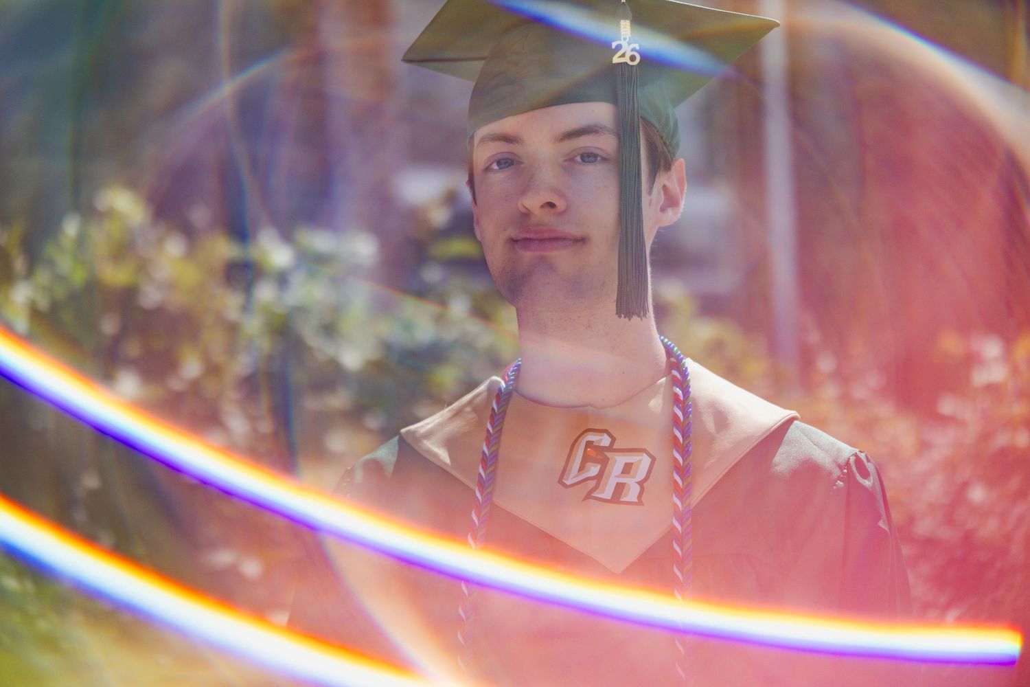 Young man in graduation cap and gown with CR insignia poses outdoors with colorful lens flare effects.