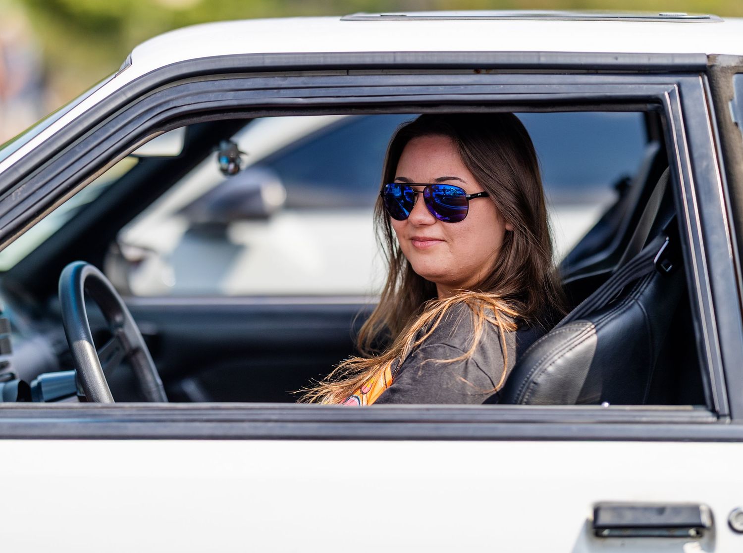 Young woman with sunglasses sitting in driver seat of white classic car, smiling confidently.