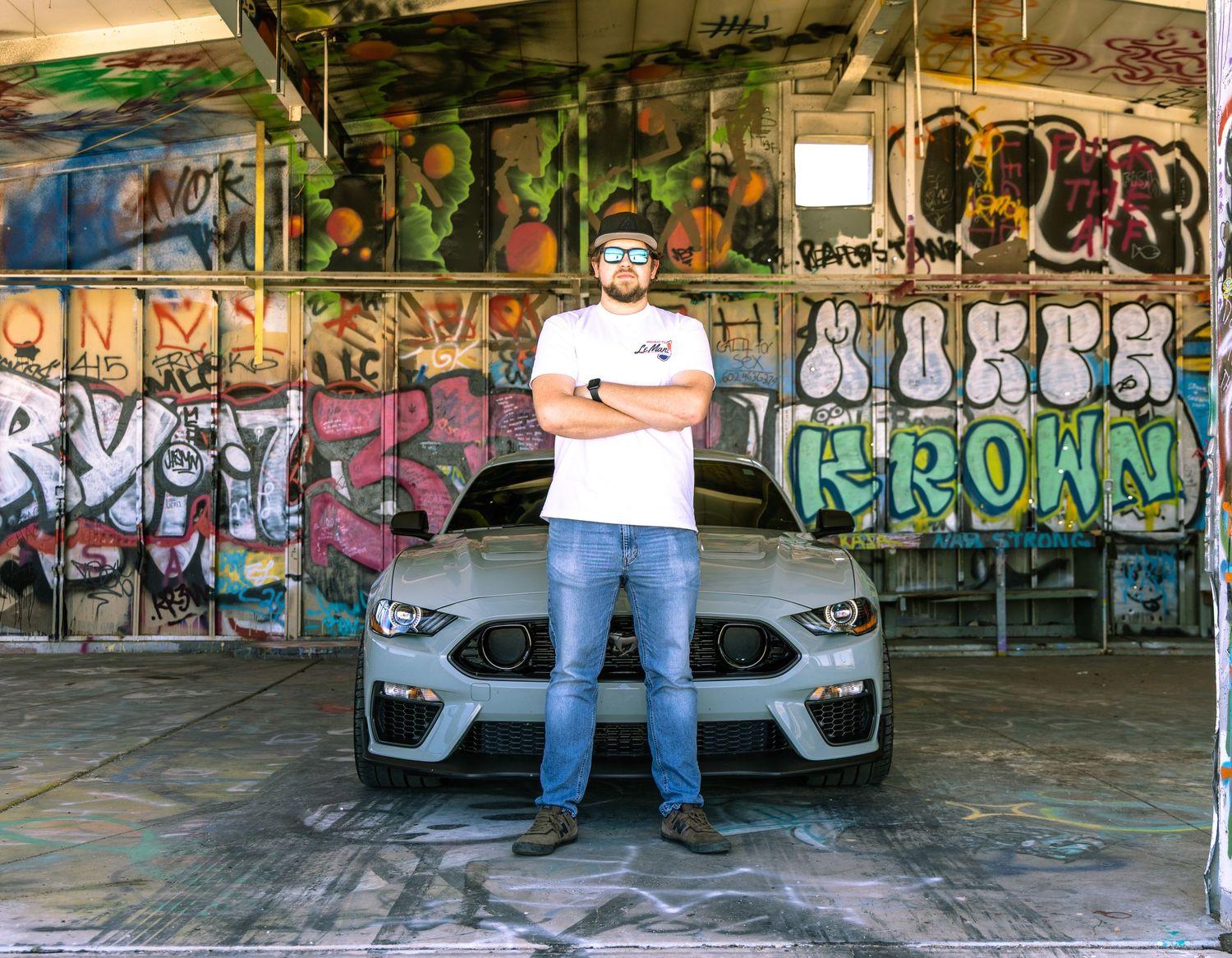 Man with crossed arms stands before a gray Ford Mustang in a graffiti-covered urban garage.