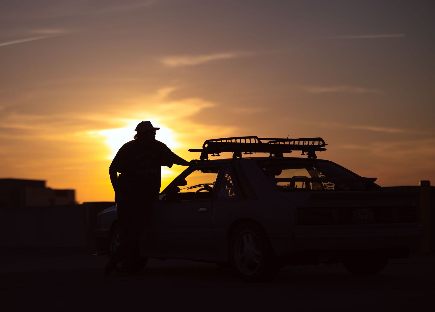 Silhouette of a person standing next to a car with a roof rack at sunset.