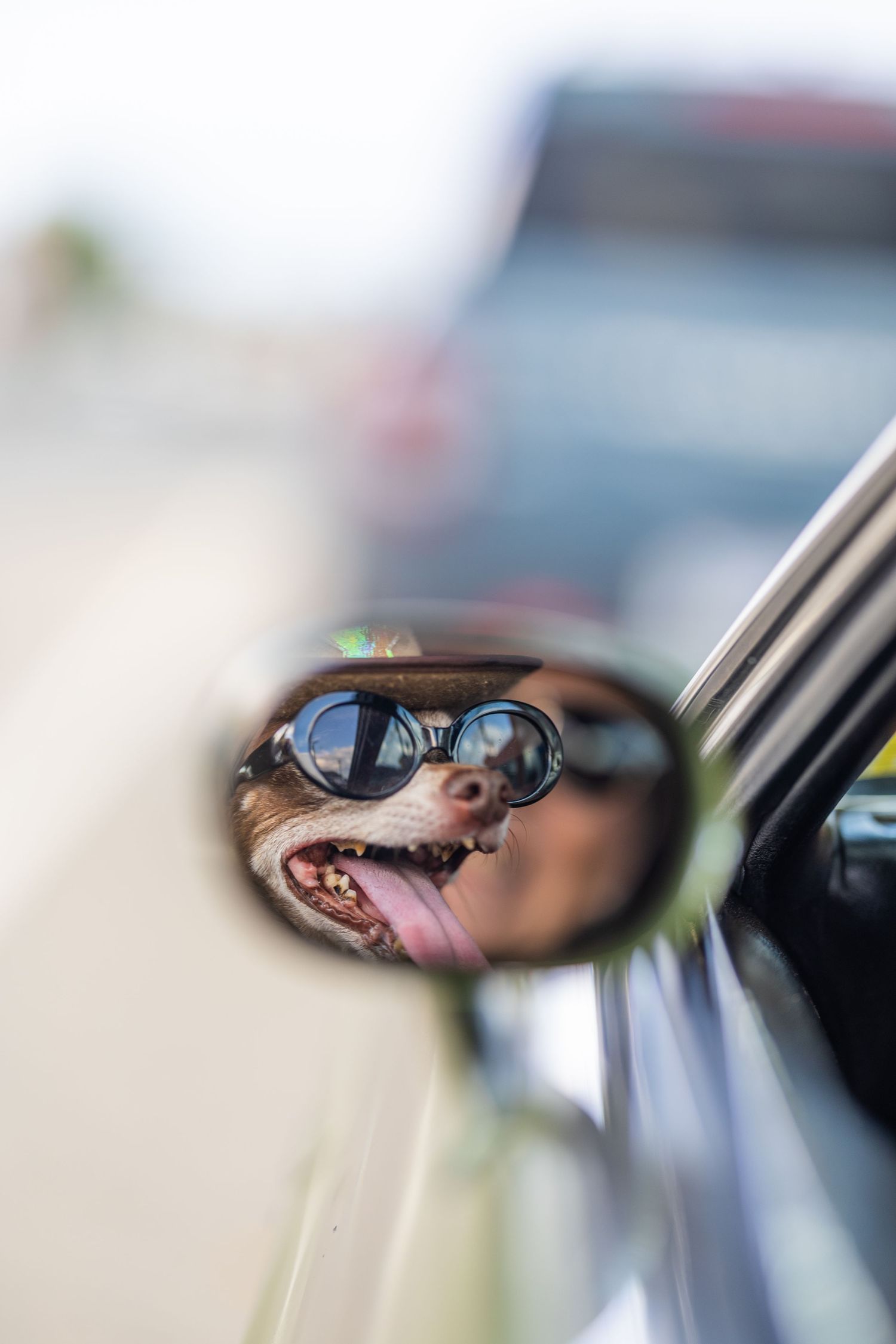 A reflection in a car side mirror shows a driver wearing sunglasses making a silly face with tongue sticking out.