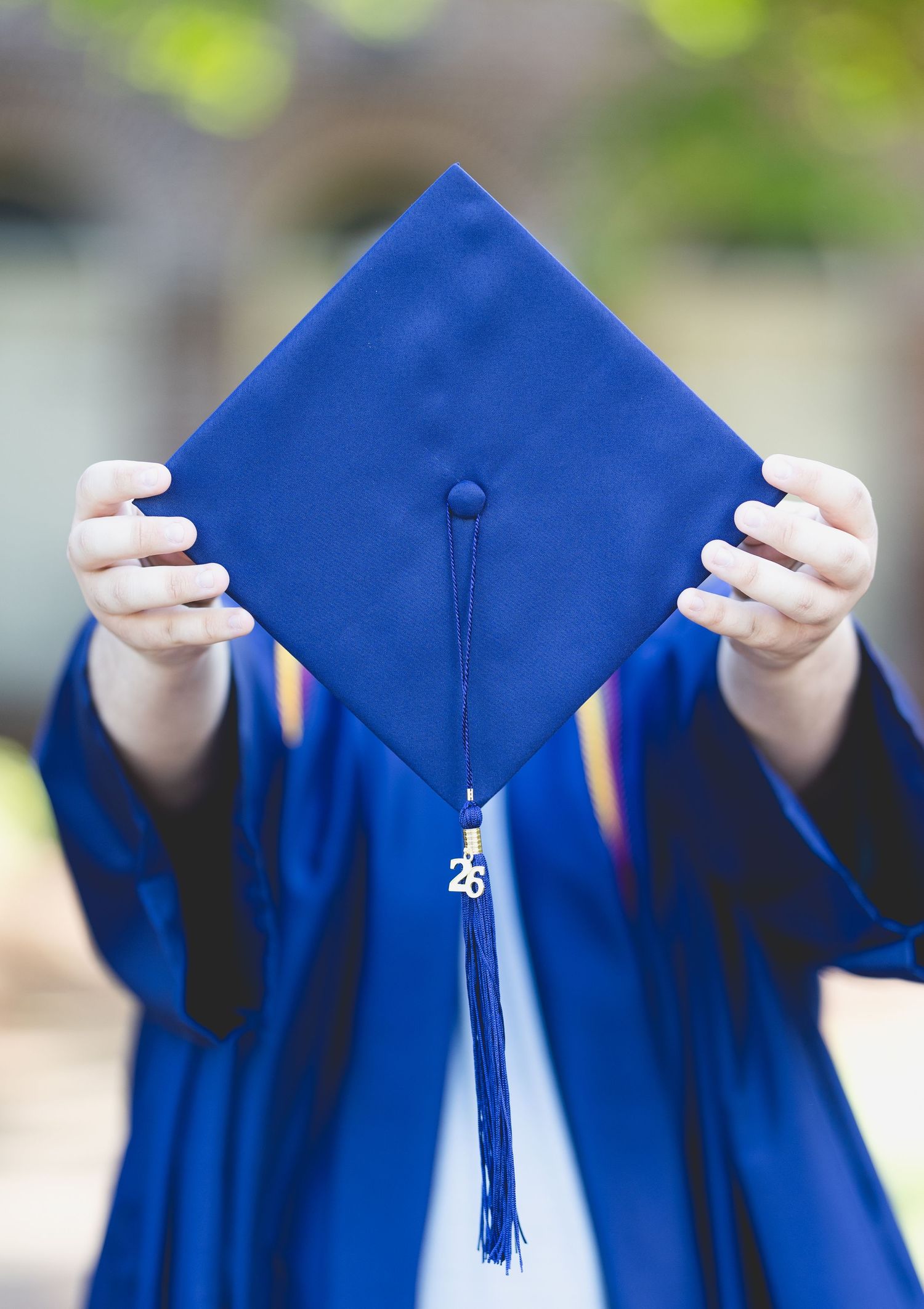 A graduate in blue cap and gown holds mortarboard up displaying tassel and 26 charm outdoors.