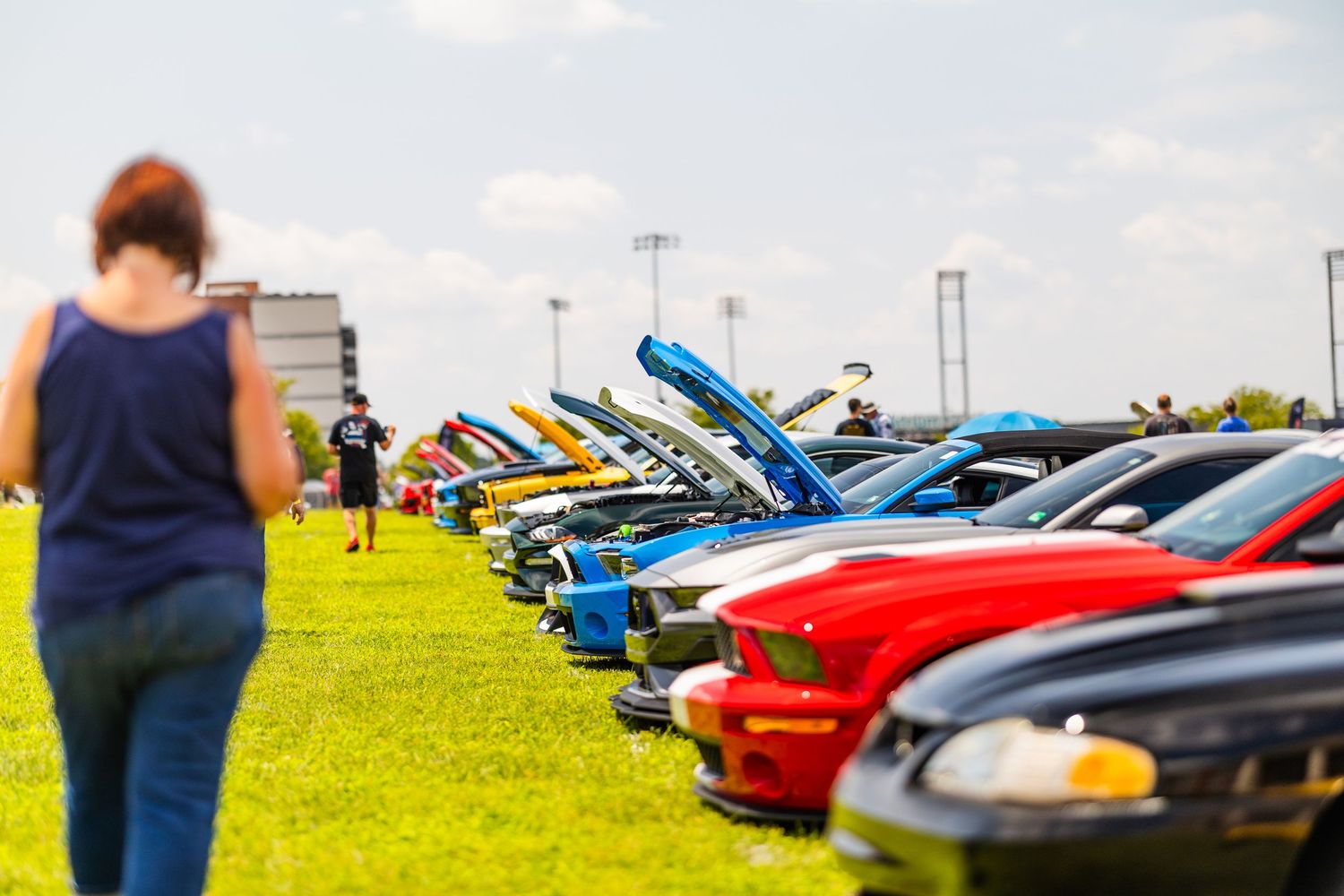 People browsing colorful classic cars with open hoods at an outdoor car show on a sunny day.
