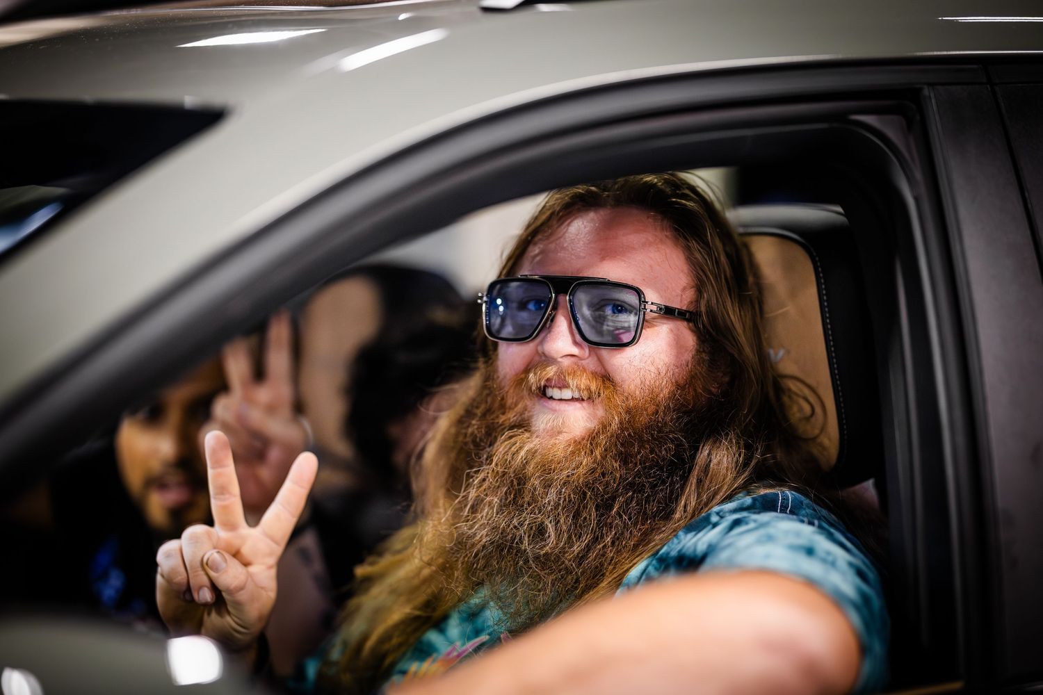 Person with beard sitting in car making peace sign gesture.
