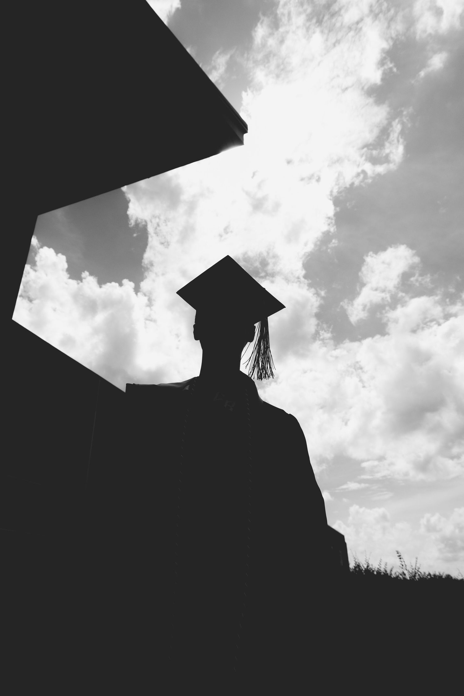 Black and white silhouette of a graduate in cap and gown against a dramatic cloudy sky.