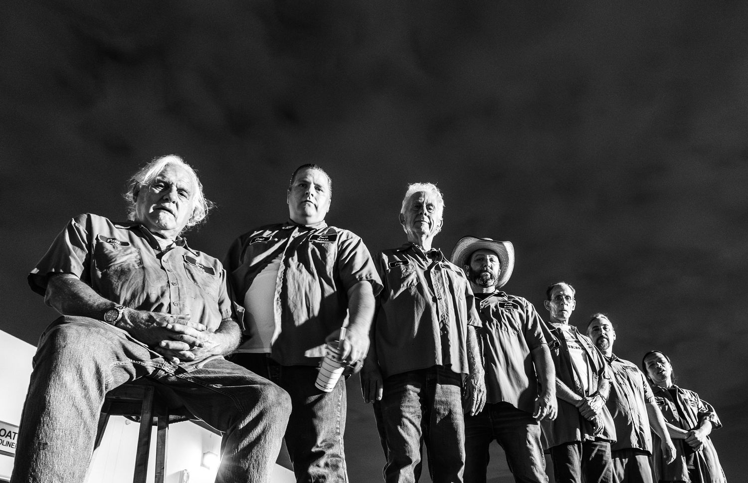 A black and white low angle photograph shows several people standing in a row against a dark background.