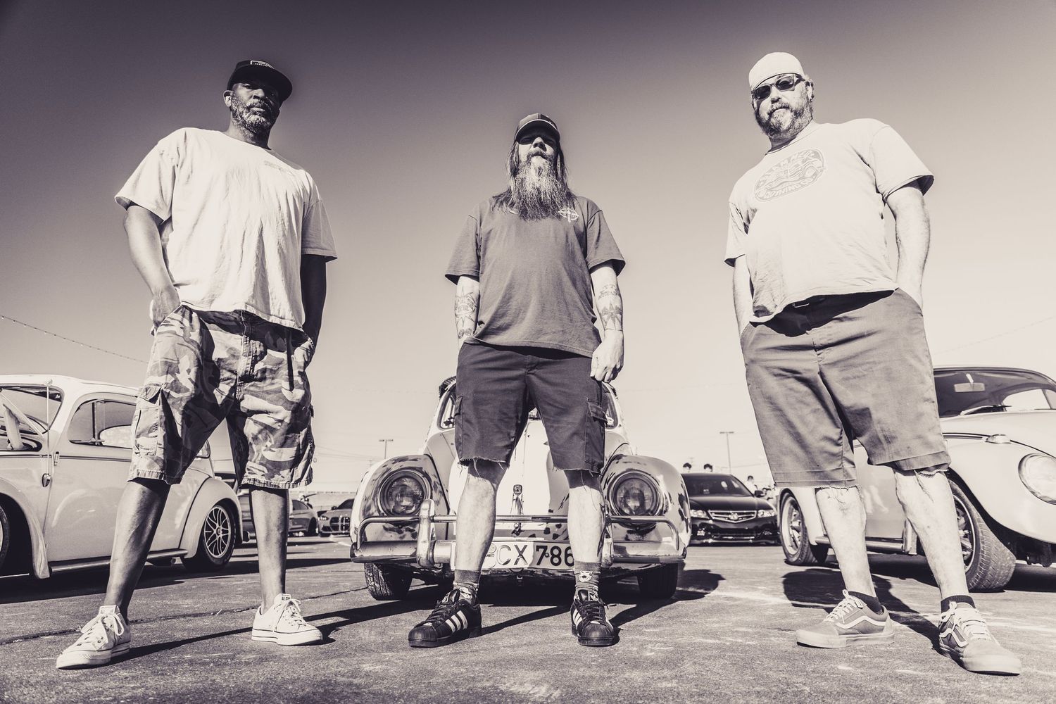 Three men posing outdoors in black and white photo with vintage cars in background at a car show.