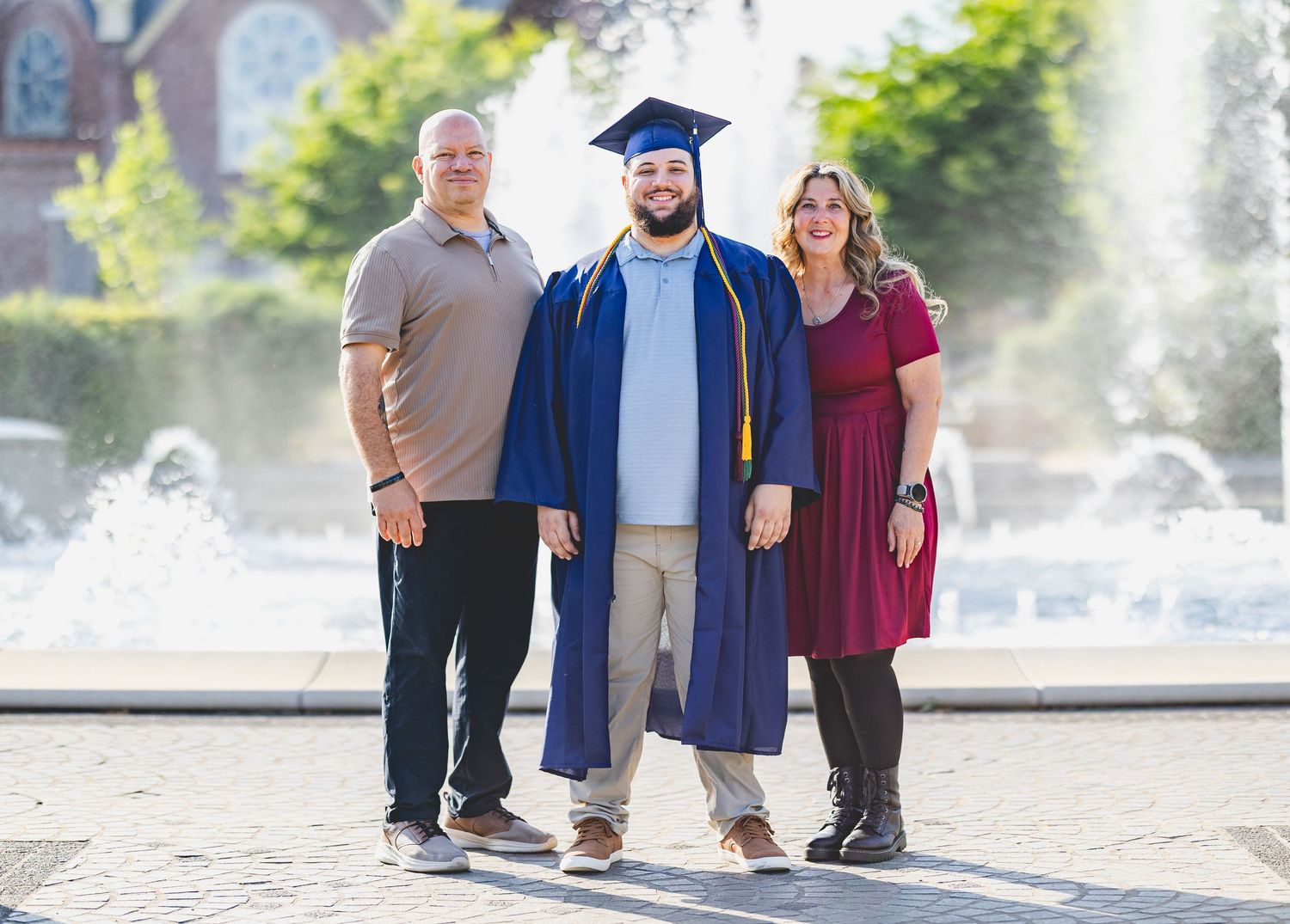 Graduate in blue cap and gown poses with proud parents in front of a fountain on college campus.
