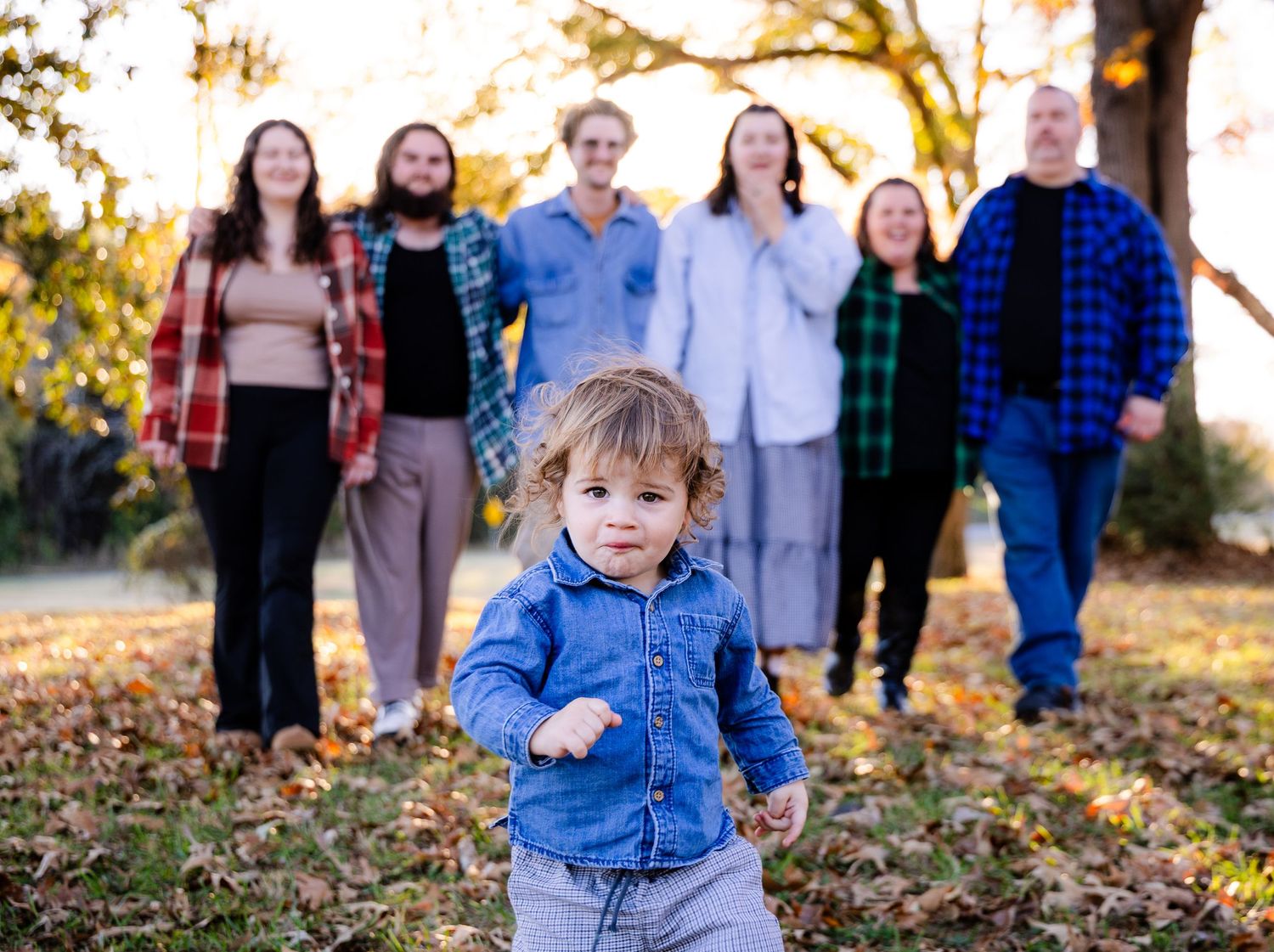 A child in blue denim runs through fallen autumn leaves during a family photo session at sunset.
