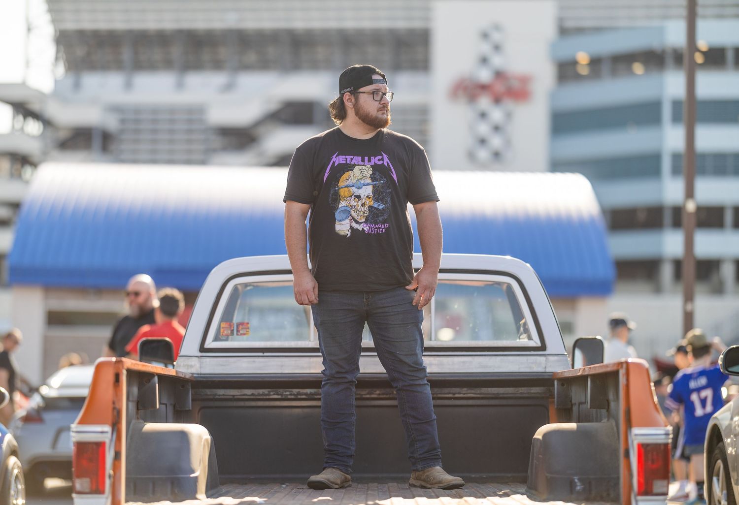 Man in Metallica t-shirt standing in truck bed at outdoor NASCAR racing event.
