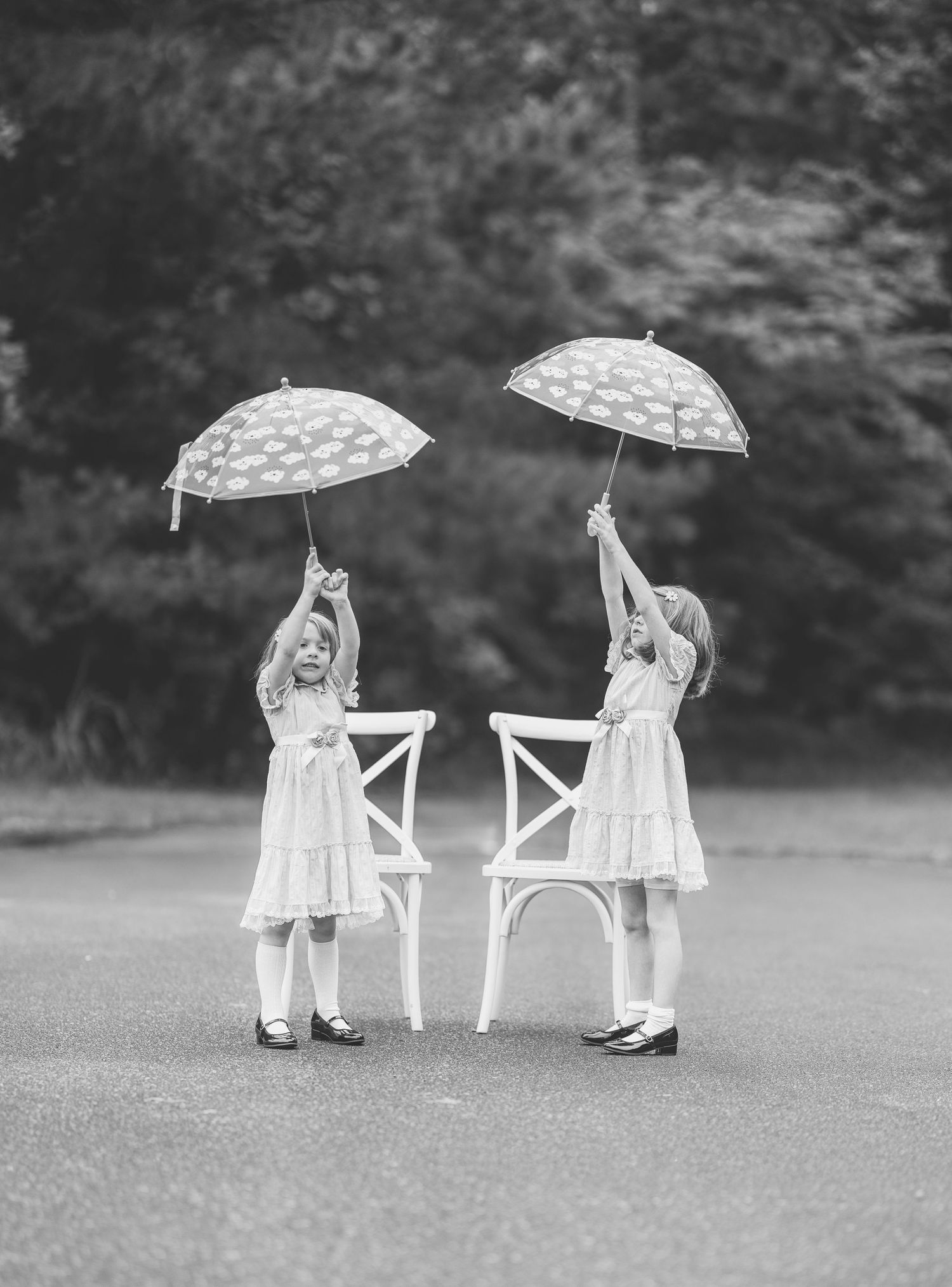 Two young girls in white dresses holding polka dot umbrellas beside a white chair in a black and white photo.