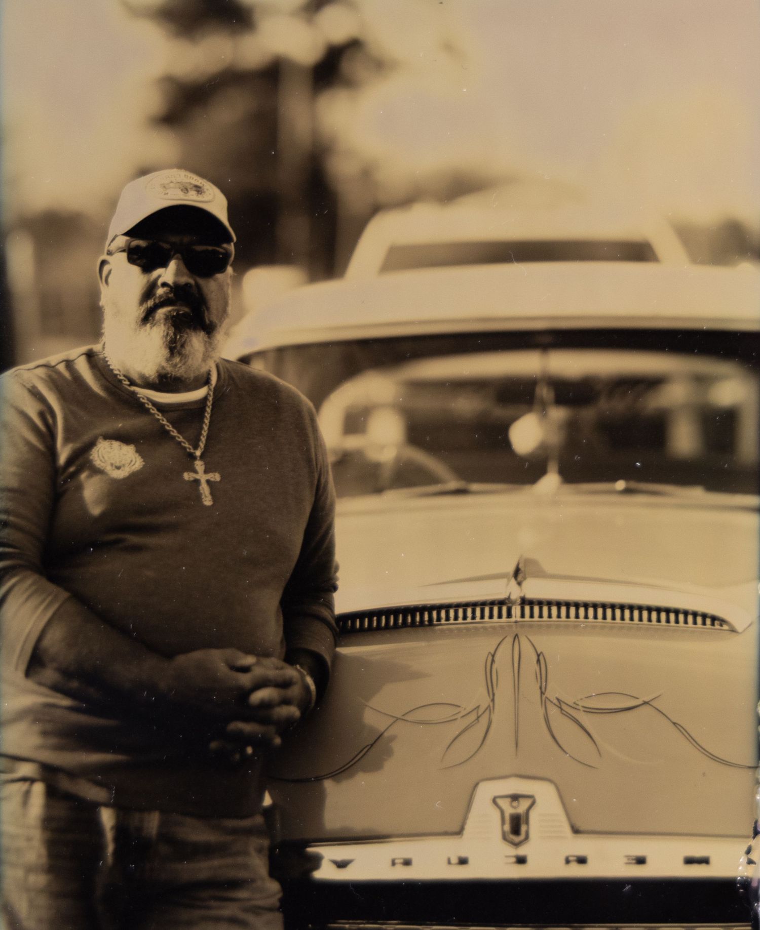 Man with sunglasses and cross necklace leaning against a vintage classic car in a sepia-toned outdoor photo.