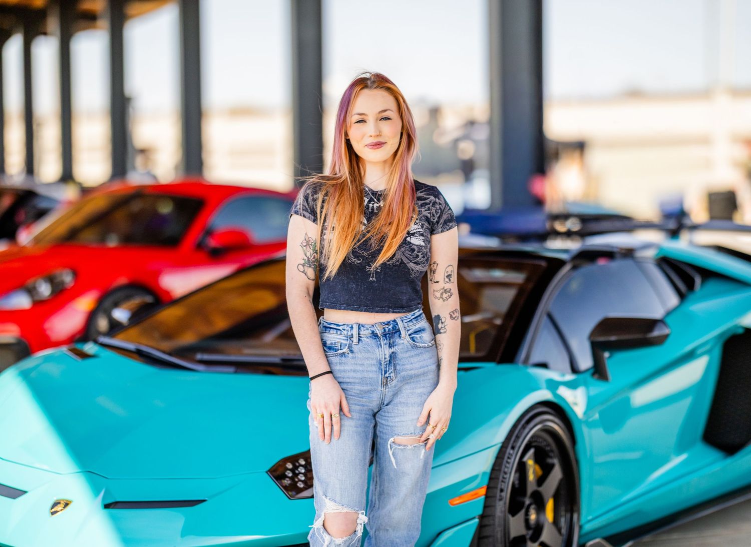 Woman with red hair and tattoos poses in front of a teal Lamborghini Aventador at a luxury car dealership.
