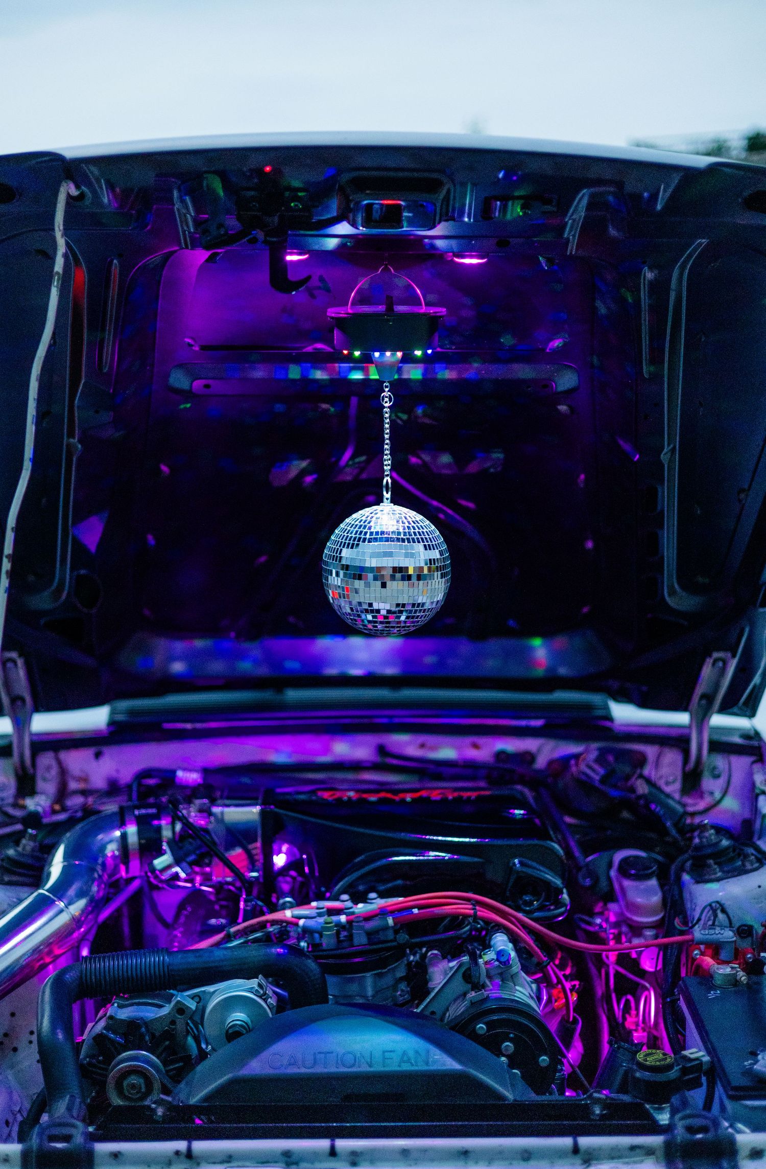 Car engine bay decorated with a hanging disco mirror ball and purple LED lights for a unique custom show car display.