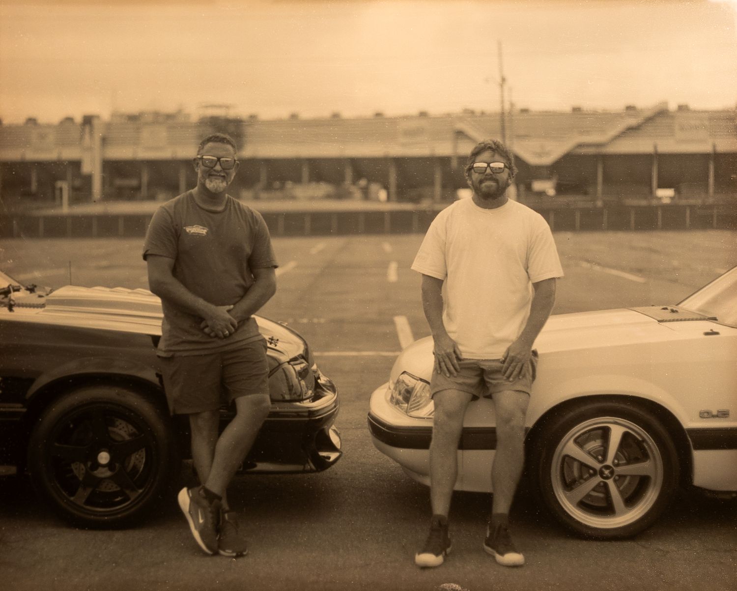 Two men in sunglasses pose between classic convertible cars in a vintage sepia-toned photo at a parking lot.