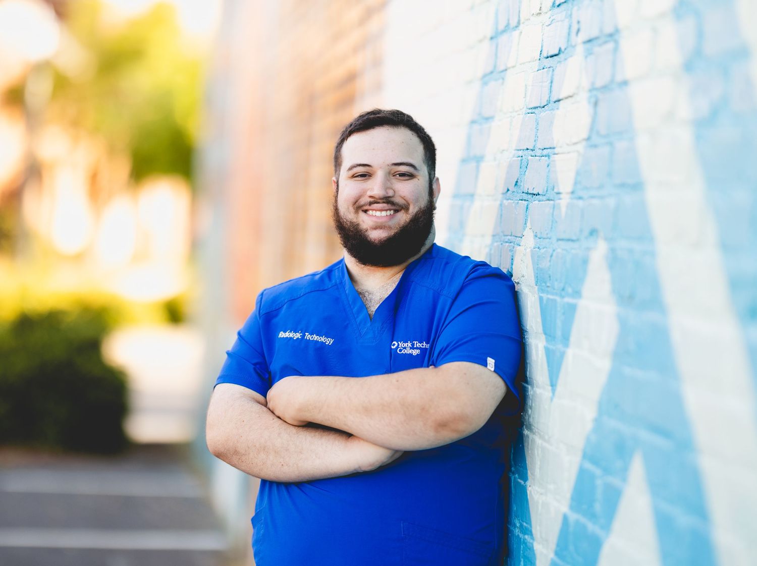 Smiling male healthcare professional in blue scrubs leaning against colorful mural wall outdoors.