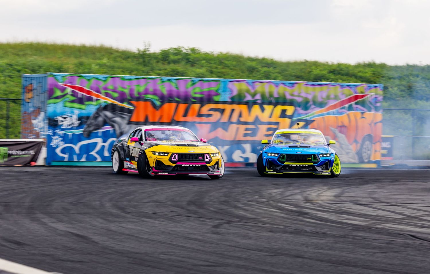 Two drift cars compete on a track at Mustang Week, with colorful graffiti wall backdrop.