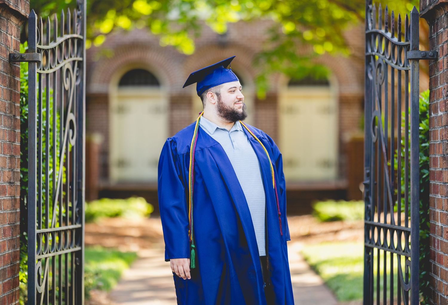 College graduate in blue cap and gown standing at ornate iron campus gates.