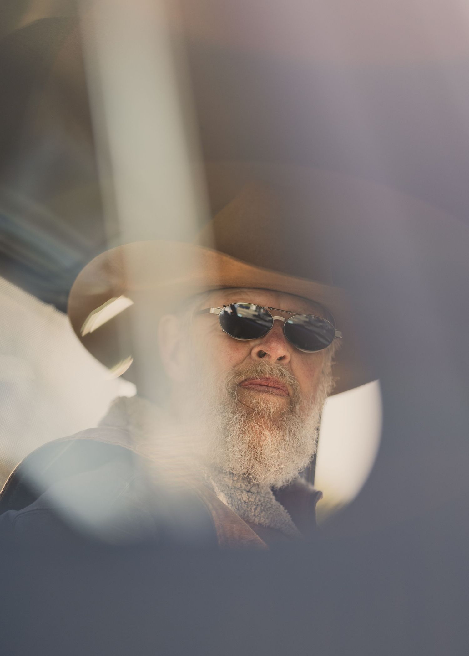 Bearded man wearing sunglasses and a wide-brim hat viewed through a car window in soft natural light.