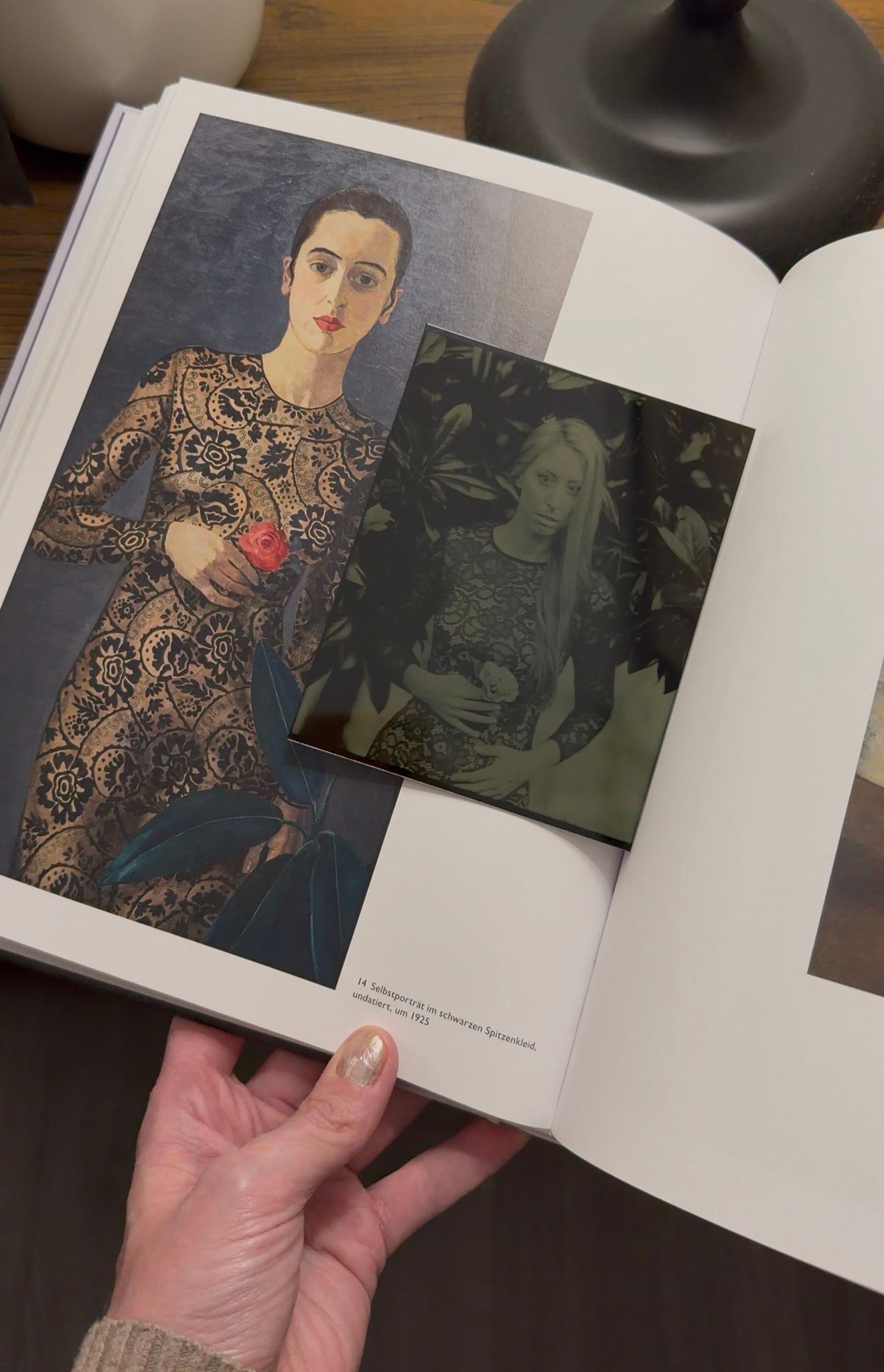 Hand holding open art book showing painting of woman in lace dress beside dark photo, on wooden table.