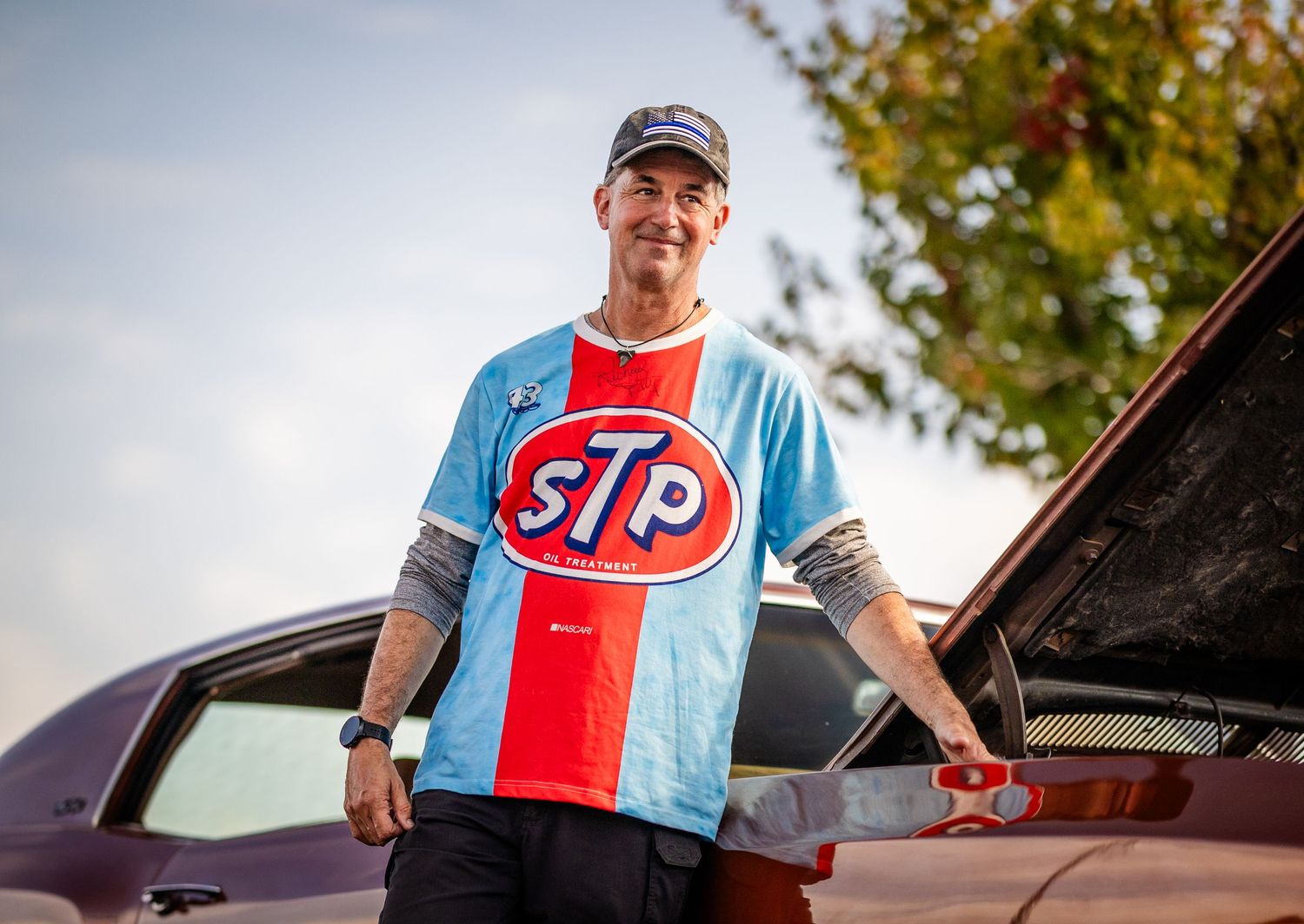 A person wearing a blue and red STP racing shirt works on a car engine under an open hood.