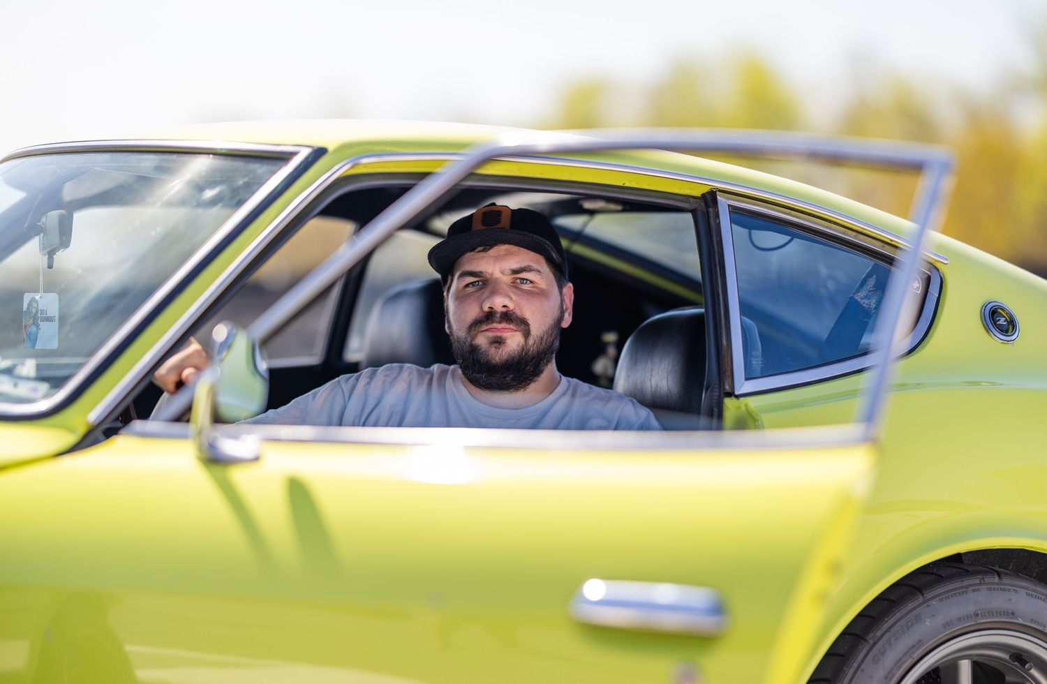 Man with beard sitting in a vintage lime green classic car, smiling through open window on sunny day.