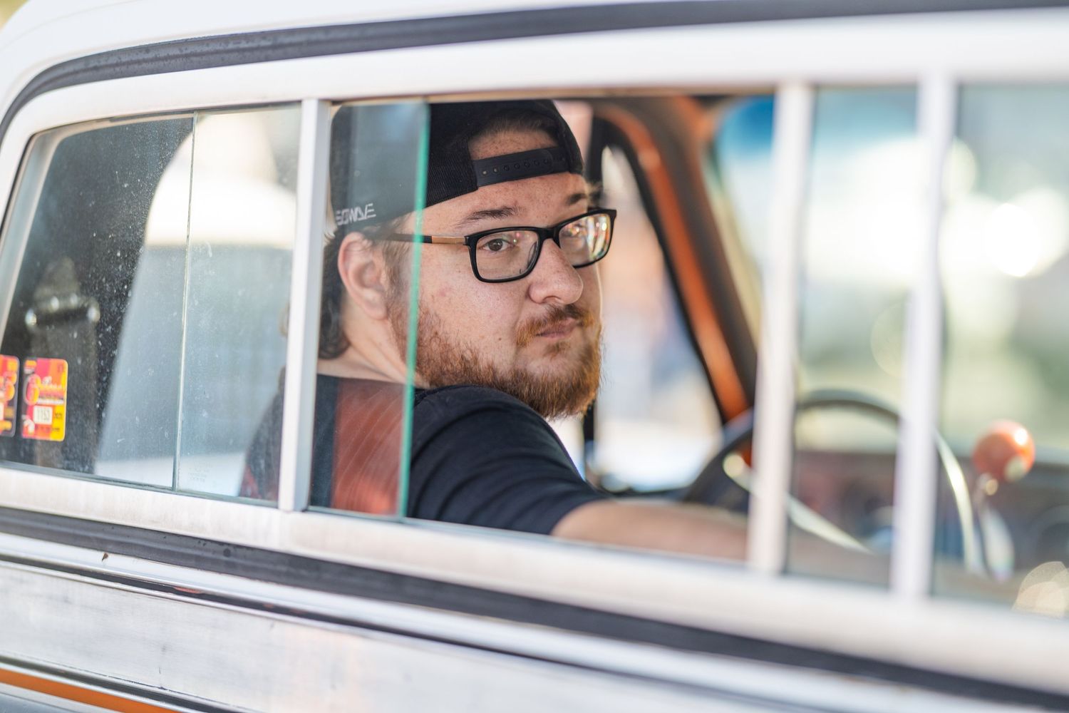 Bearded man with glasses and backward cap looking back through vintage car window.