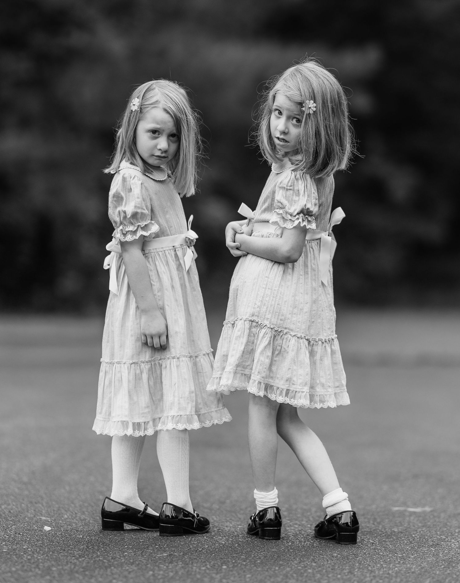 Two young girls in matching vintage white dresses and black shoes pose outdoors in black and white photo.