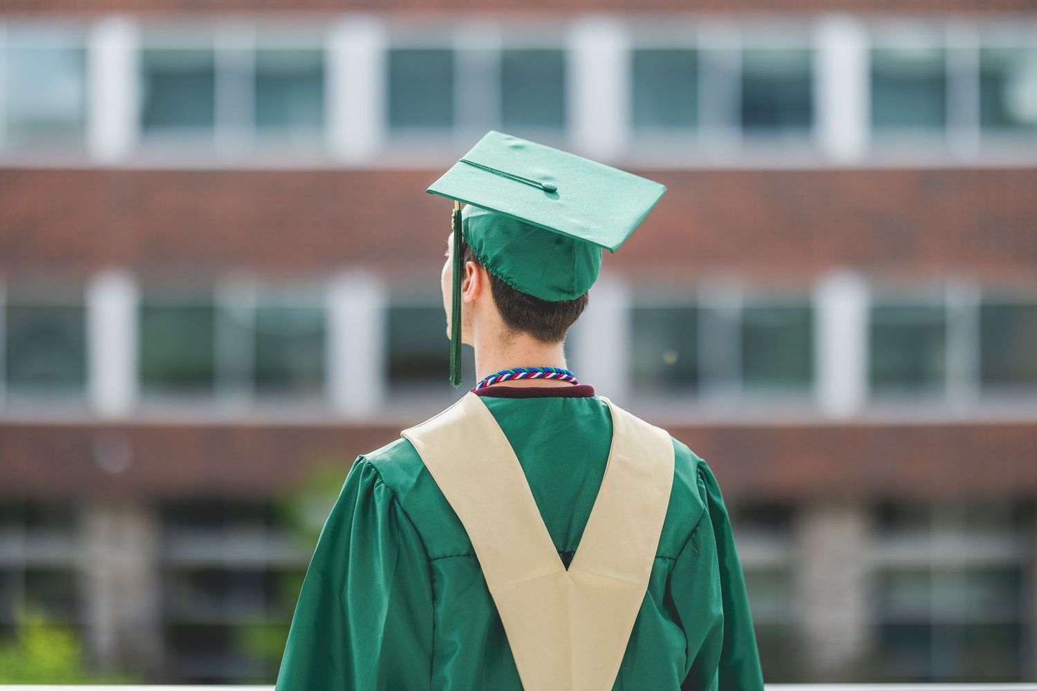 Graduate in green cap and gown stands facing a brick university building on graduation day.