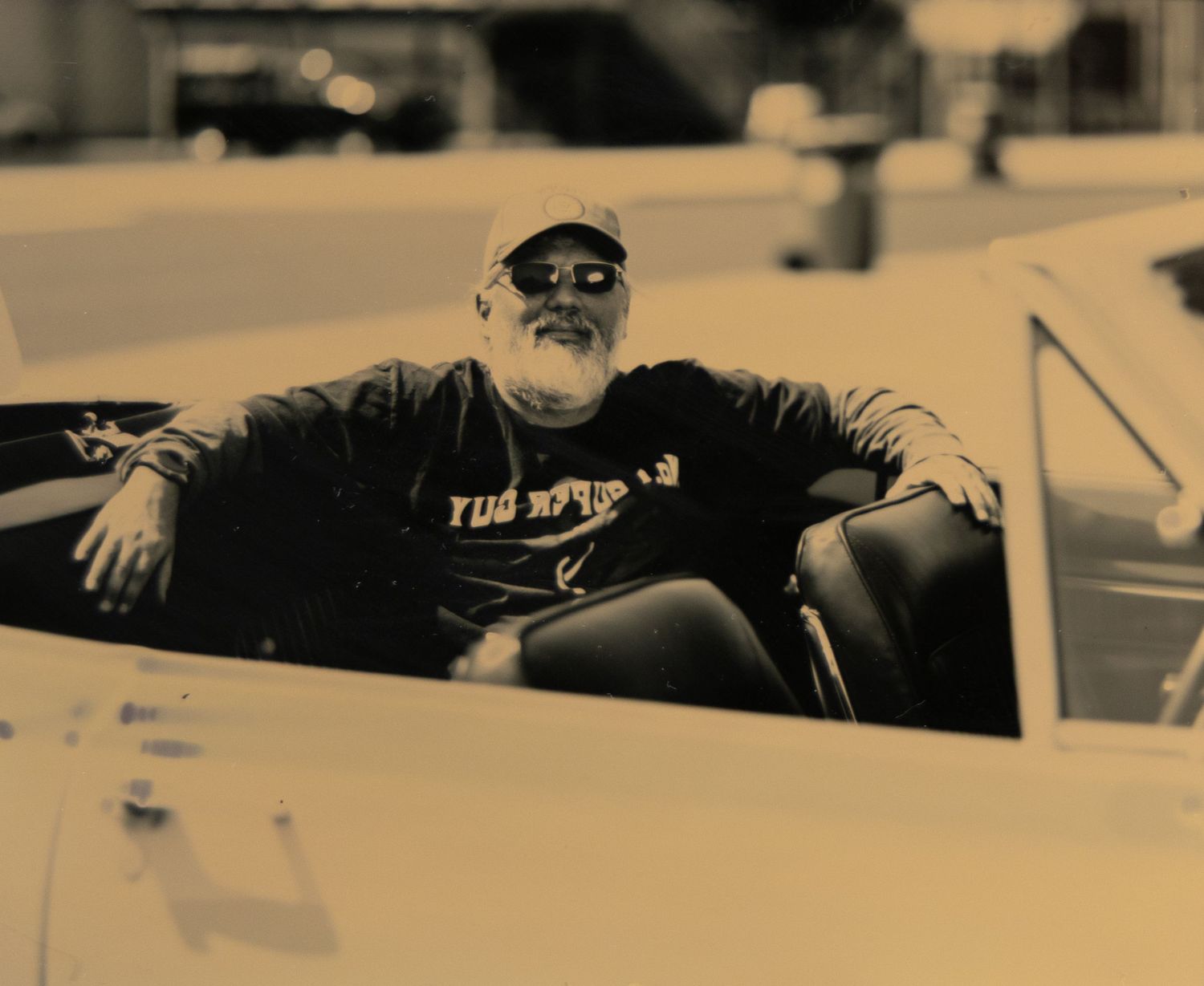 Older bearded man relaxing in a vintage convertible car, wearing sunglasses and a black t-shirt, sepia-toned photo.
