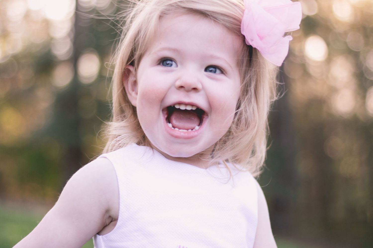 Happy toddler girl with blonde hair and pink bow laughing outdoors in soft light.