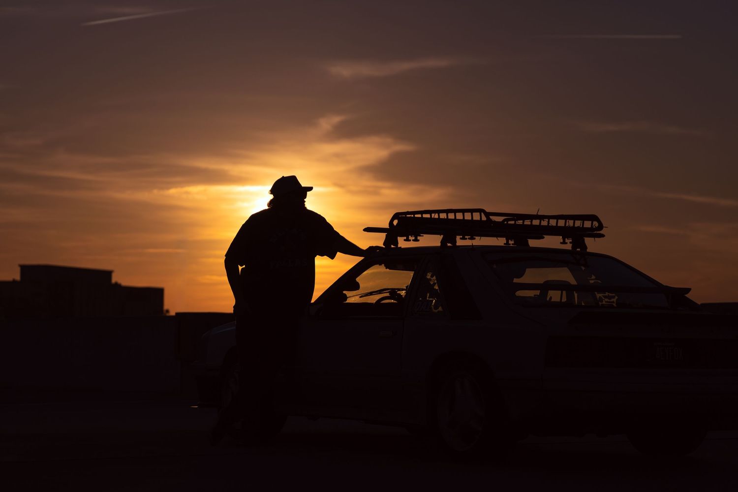 Silhouette of a man in a cowboy hat leaning on a vehicle at sunset with warm orange sky.