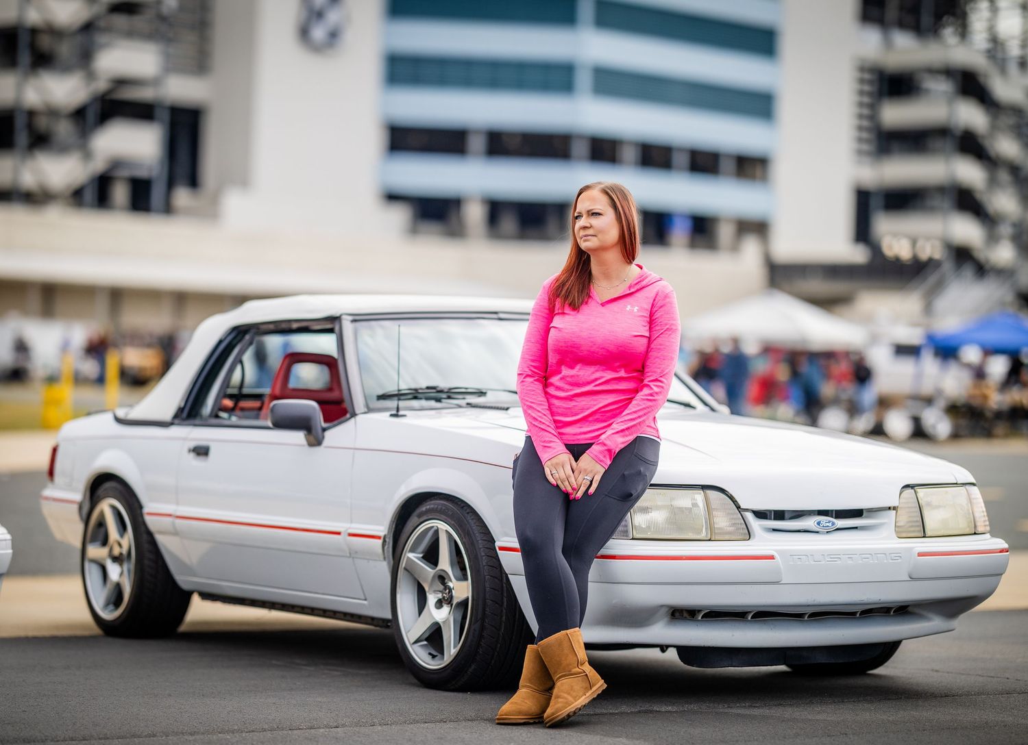 Woman in pink shirt posing with white classic Mustang at car show.
