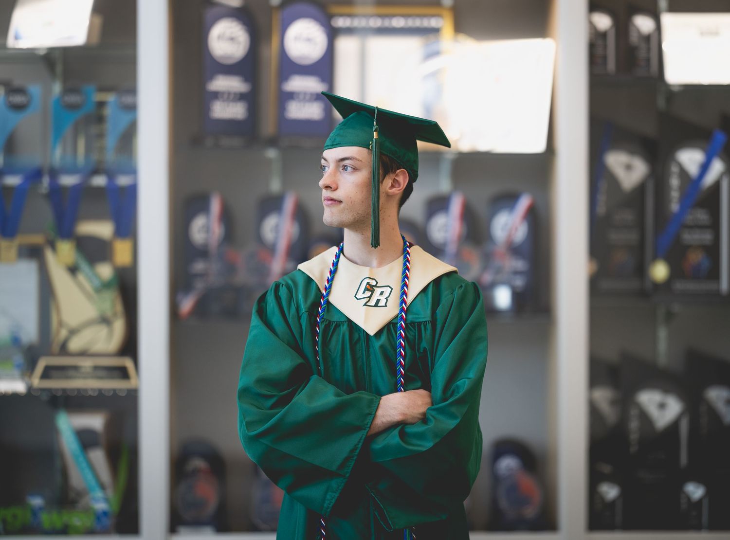 High school graduate in green cap and gown stands with arms crossed, posing in front of trophy display case.