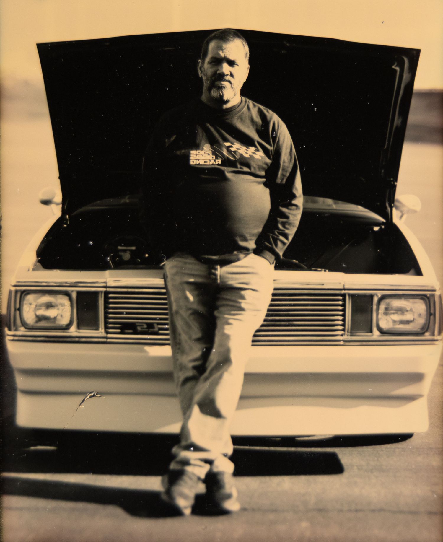 Man standing in front of classic car with open hood in sepia-toned photo.
