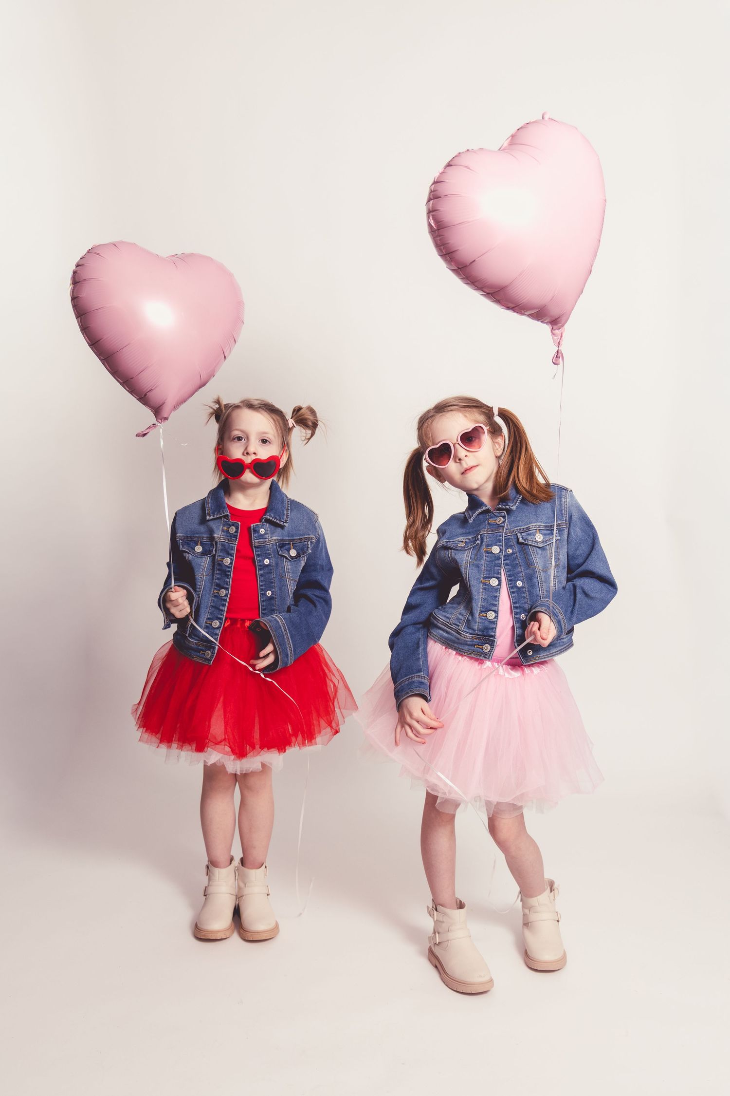 Two children in denim jackets and tulle skirts pose with pink heart balloons against a white background.