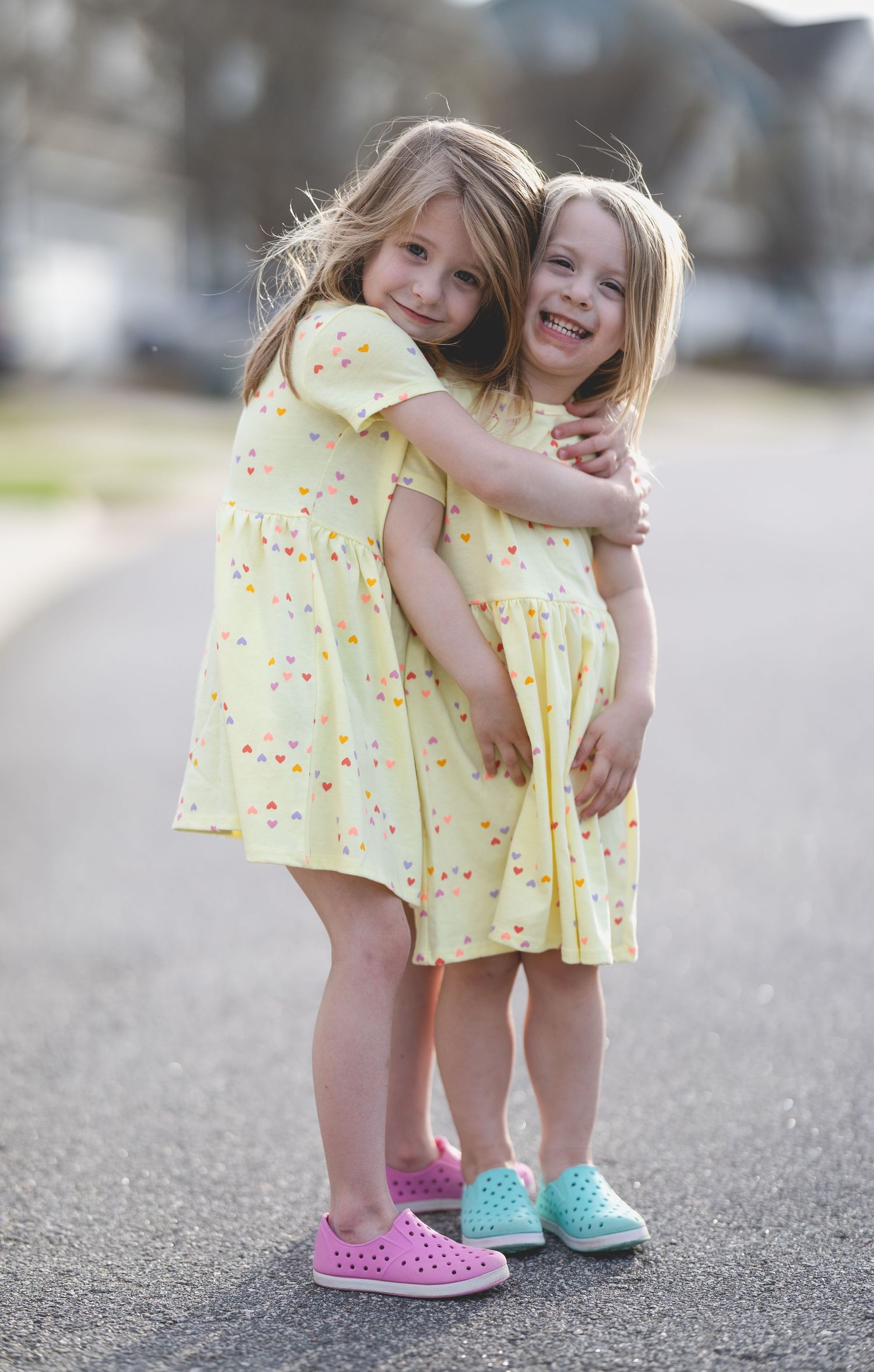 Two little girls in matching yellow dresses hugging on a street, wearing colorful shoes.