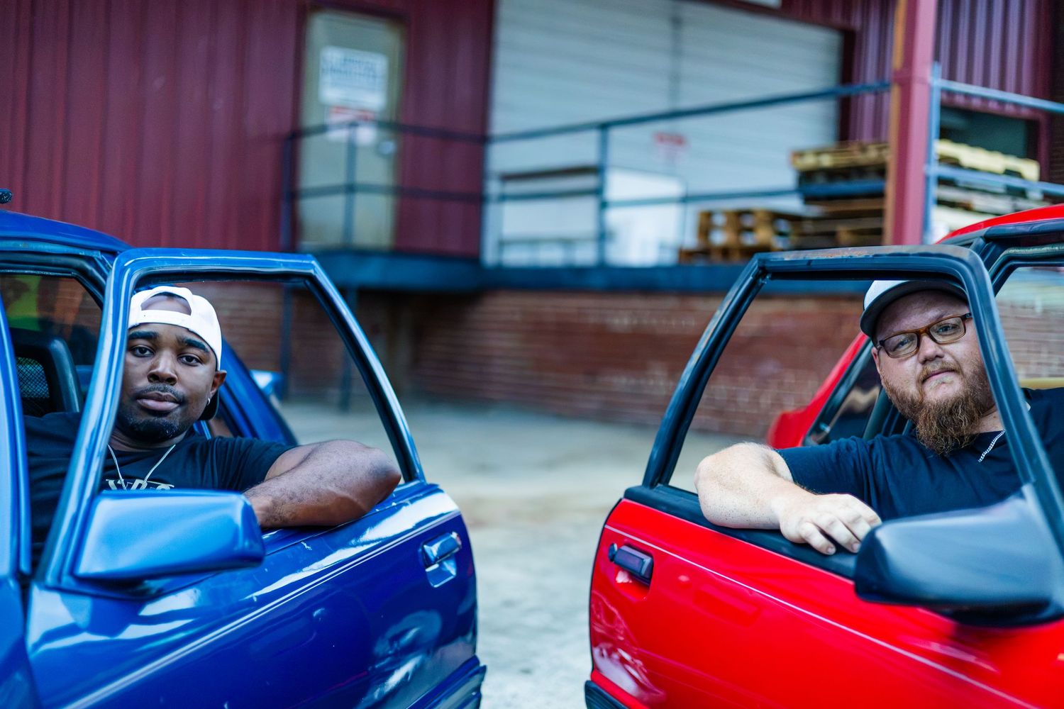 Two men leaning out of open doors of blue and red trucks parked near a red building.