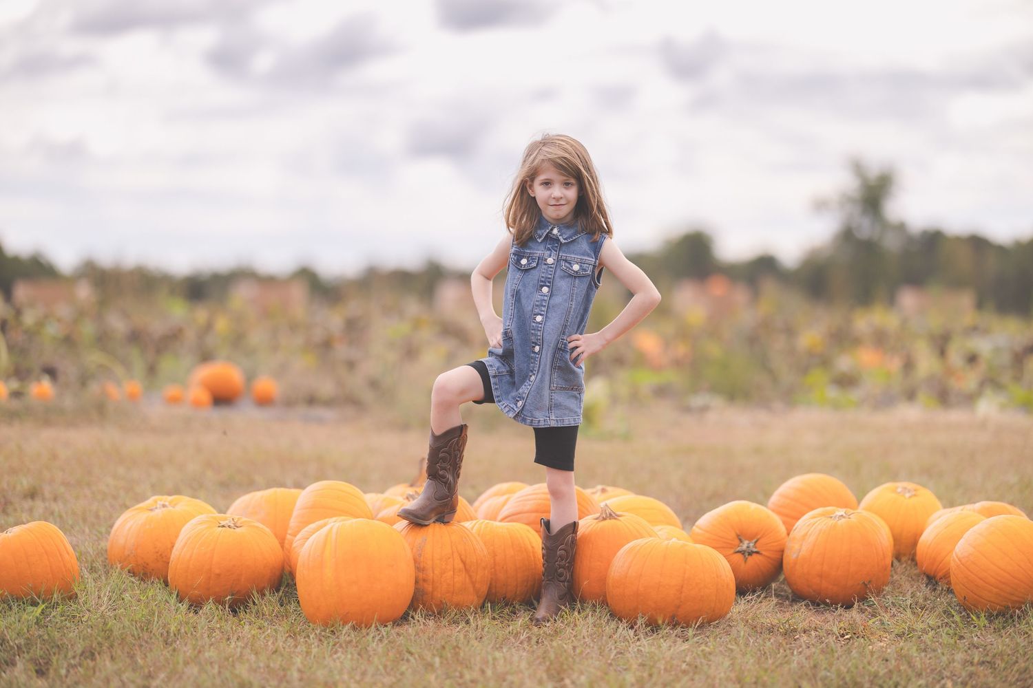 Young girl in denim dress and cowboy boots posing confidently among orange pumpkins in a fall pumpkin patch.