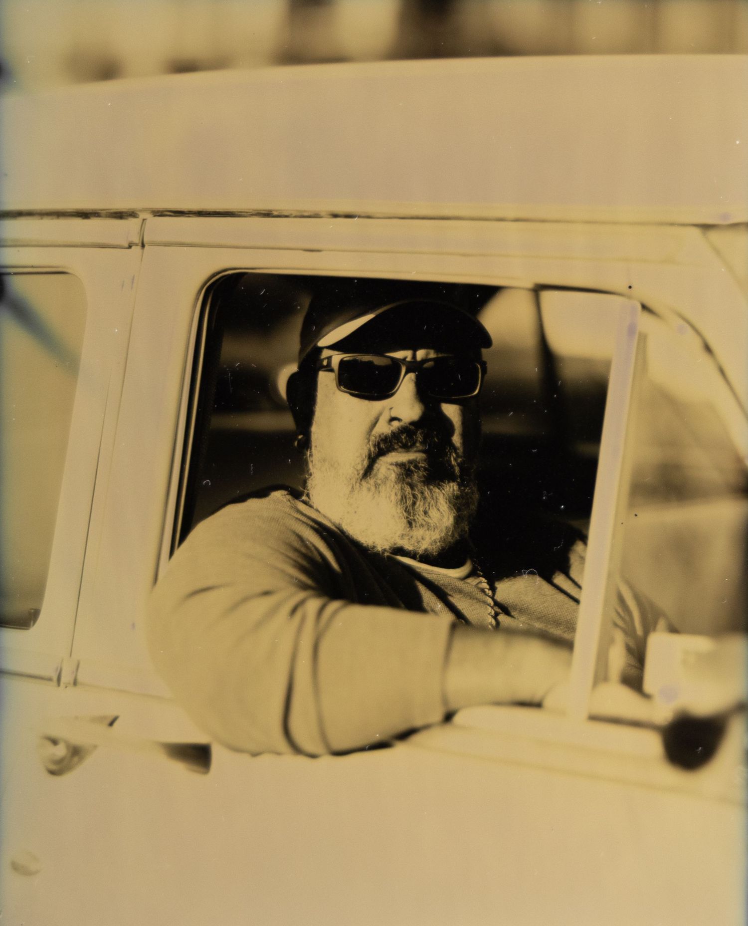 Bearded man wearing sunglasses and cap sitting in a vintage white vehicle window in a sepia-toned photo.