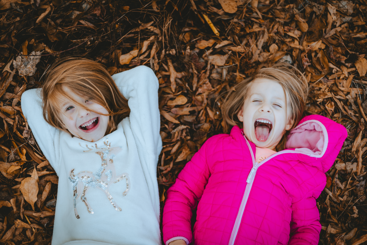 Two happy children laughing while lying on autumn leaves, wearing cozy fall outfits outdoors.
