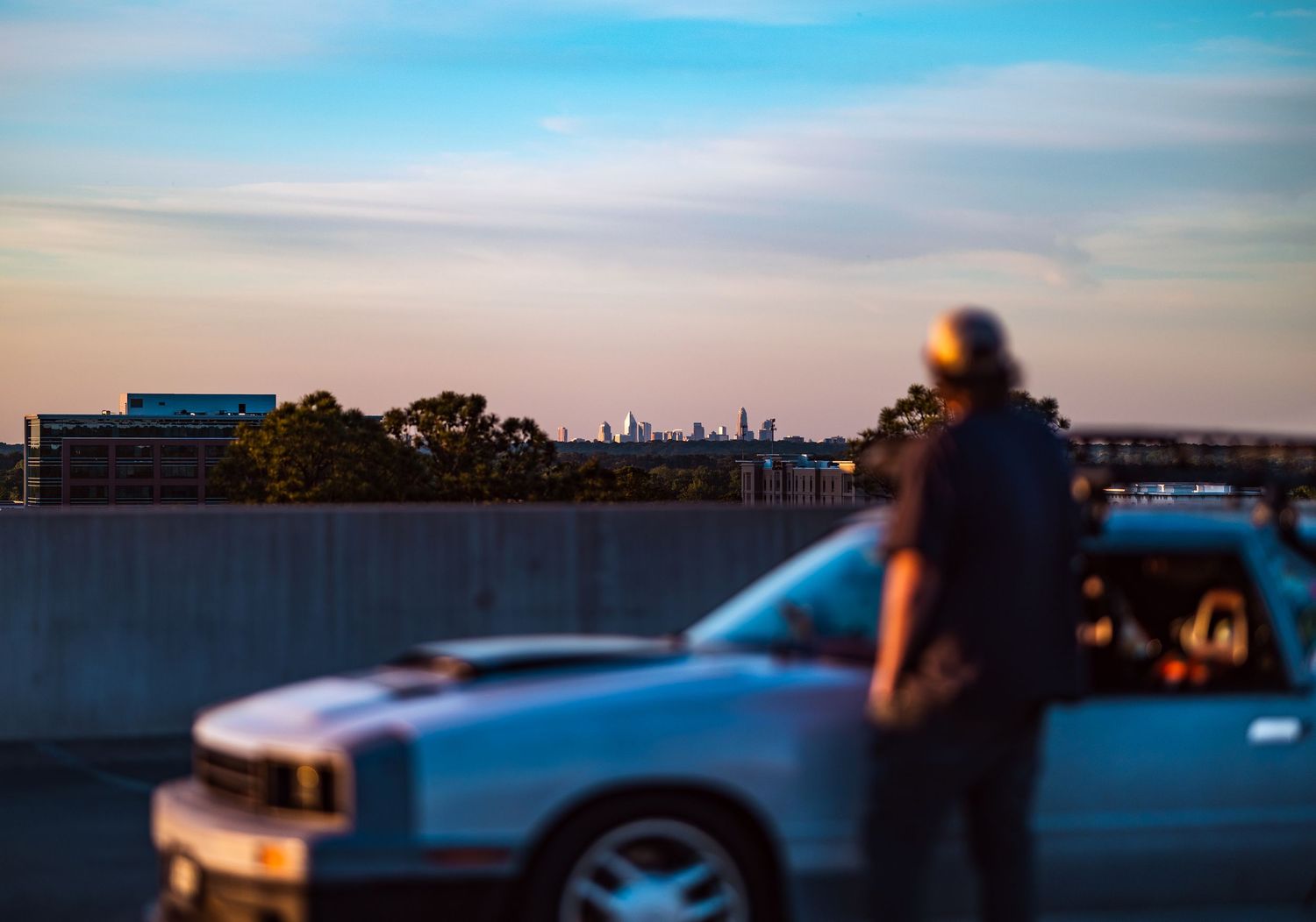 A person stands beside a silver car on an elevated road at dusk with a city skyline in the background.
