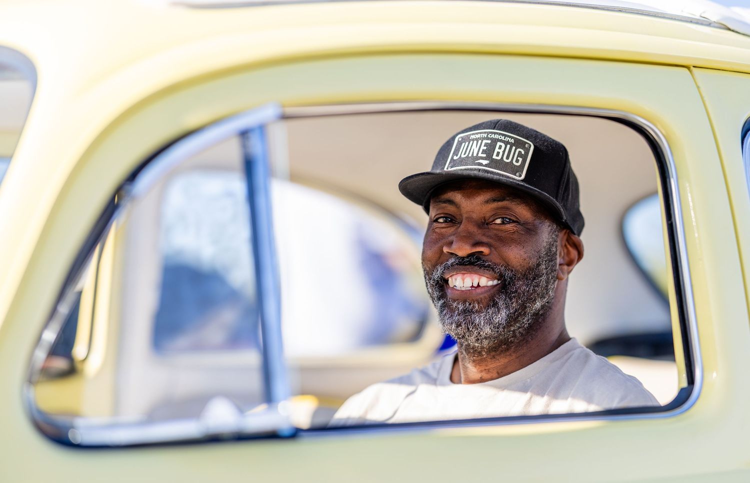 Man smiling in vintage yellow car window, wearing a black Lone Bug snapback cap outdoors.