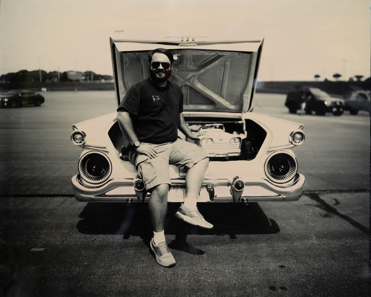 Black and white photo of a bearded man in sunglasses sitting on the trunk of a vintage classic car in a parking lot.